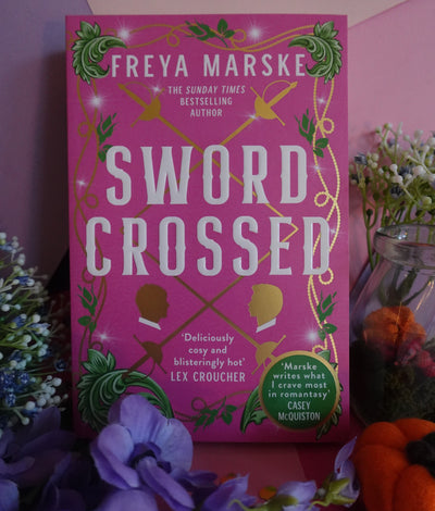 The cover of the book Swordcrossed by Freya Marske. Bright pink background, overlayed with goldd foil sillouettes of rapier swords, two faces opposite each other and otherwise ordained with stylized green laurel ornaments in the corners
